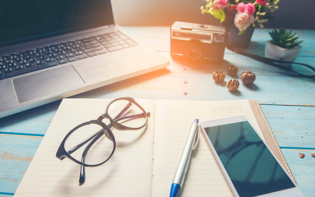 An open notebook with glasses, pen, and phone beside a laptop.
