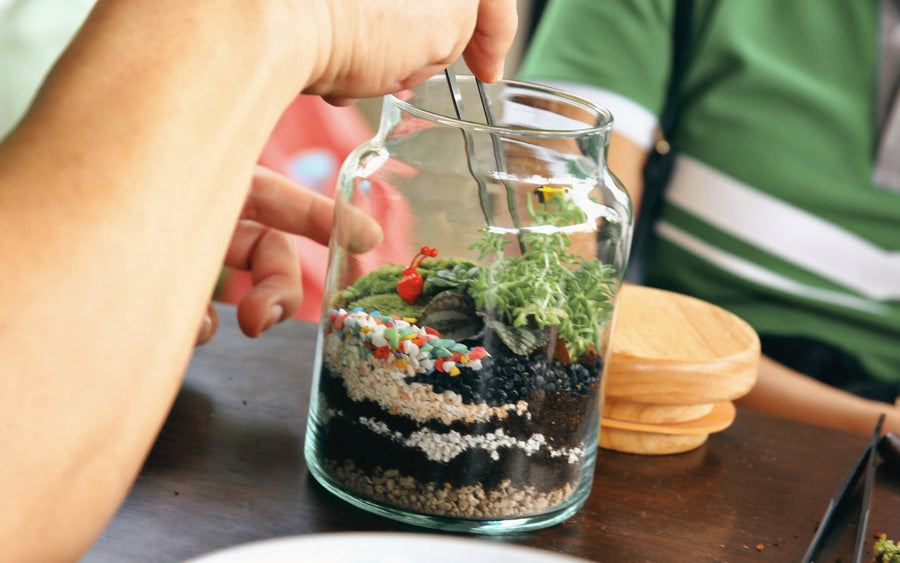 A hand uses tweezers to arrange plants inside a glass terrarium.