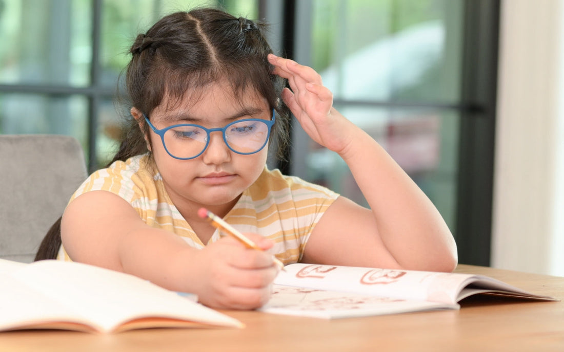A girl wearing blue glasses writes in an open workbook at a table.