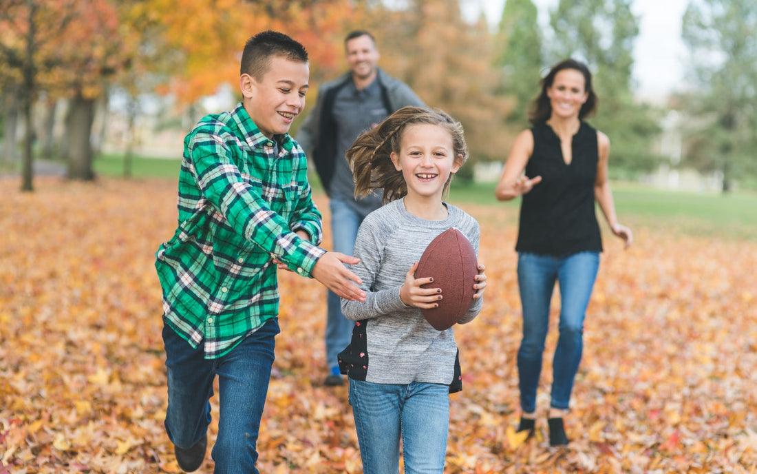 Two children carry a football through fallen leaves with adults behind them.
