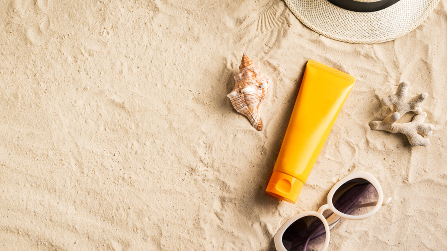 A yellow sunscreen tube, sunglasses, and seashells on sand.