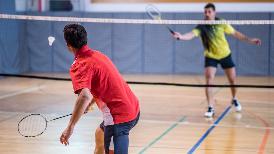 Two people play badminton across an indoor court.